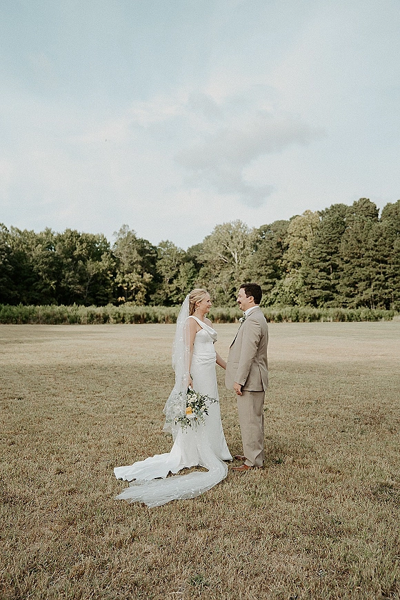 Couple portrait of bride and groom facing each other, her long veil and bouquet in an open field with tree line under cloudy sky