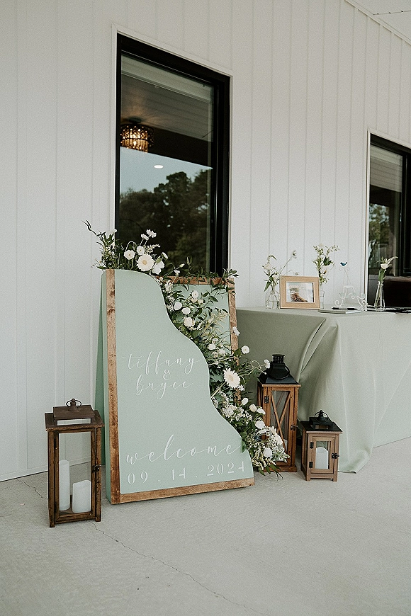 Wedding welcome sign in a wood frame with calligraphy and floral greenery swag, flanked by lanterns and candles on a patio table