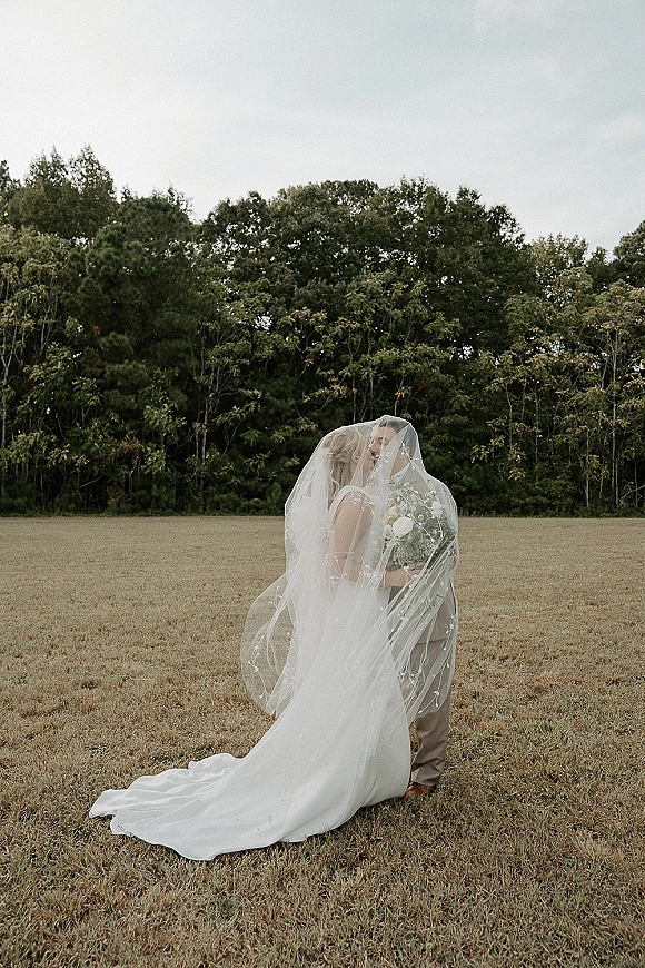 Wedding couple portrait with a long veil draped over them as they embrace, bride holding a white rose bouquet in a grassy field