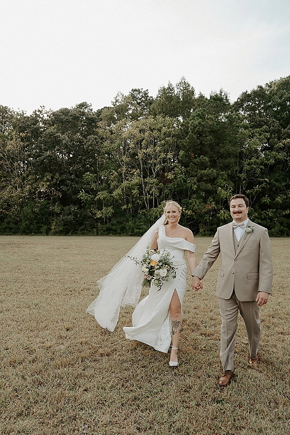 Couple portrait of bride and groom holding hands, bride with long veil and bouquet, groom in tan suit in a grassy field under open sky