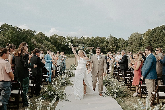 Wedding recessional as newlyweds walk down the aisle, bride lifting bouquet overhead beside groom, with veil, guests, arch, and greenery in a field