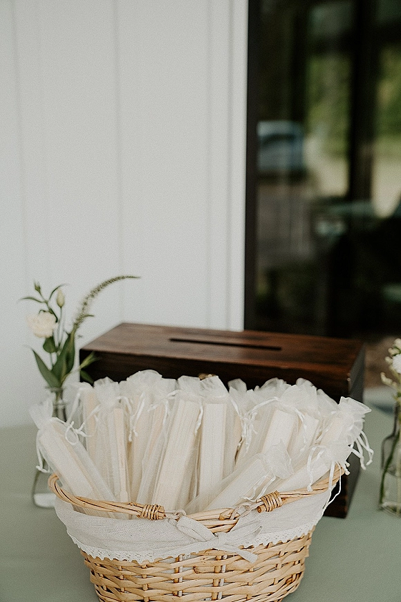 Wedding favor display with wedding favor bags in a wicker basket, lace trim and white flowers in bud vases by a window