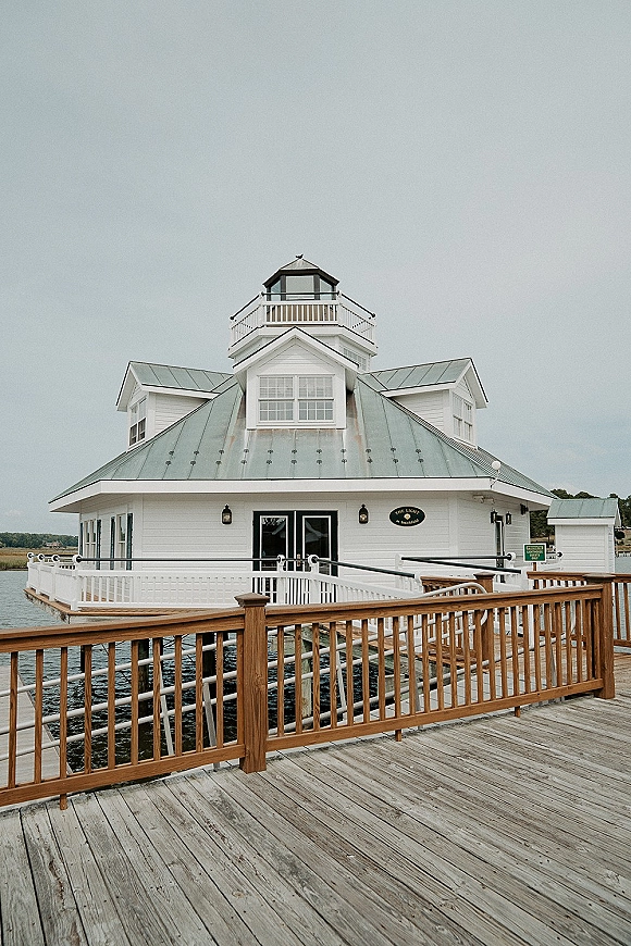 Waterfront wedding venue with a white wood building and cupola beside a dock, overlooking marsh grass shoreline under overcast sky