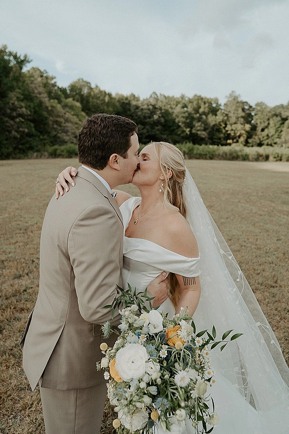Wedding kiss portrait of bride and groom kissing, her veil flowing as she holds a bouquet in an open field under cloudy skies