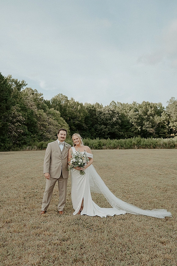 Couple portrait outdoors, bride in off-the-shoulder dress holding a bouquet beside groom in tan suit with bow tie in a grassy field