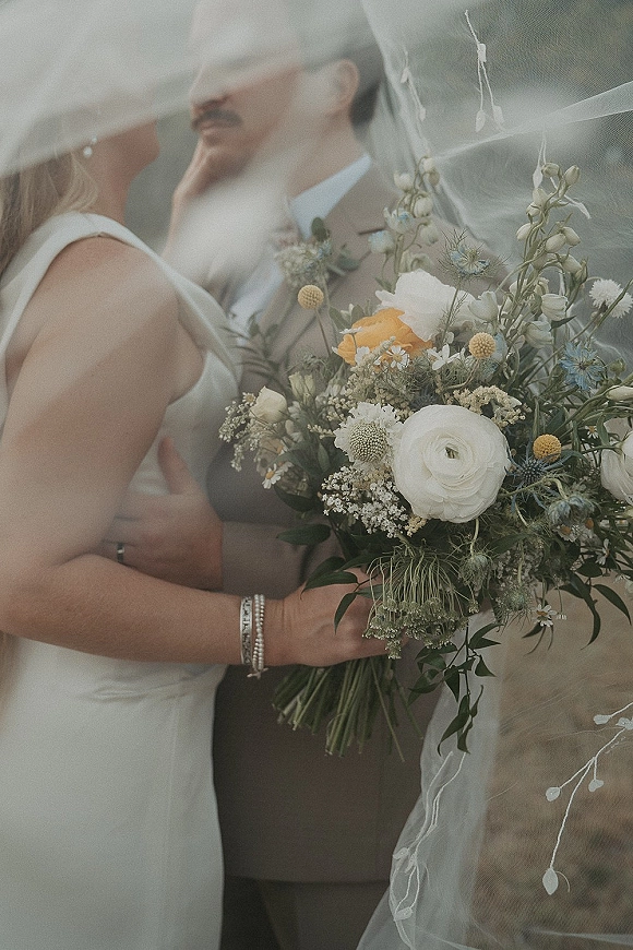 Wedding couple portrait of bride and groom embracing under a veil, bride holding a wildflower bouquet with outdoor greenery behind