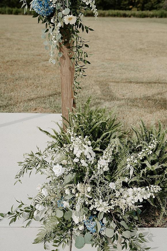 Wedding ceremony flowers with blue hydrangea and white blooms, ferns and eucalyptus on a wood post, set on a grassy outdoor field