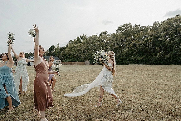 Bouquet toss as the bride throws her wedding bouquet over her shoulder, long veil flowing toward cheering bridesmaids in an open field