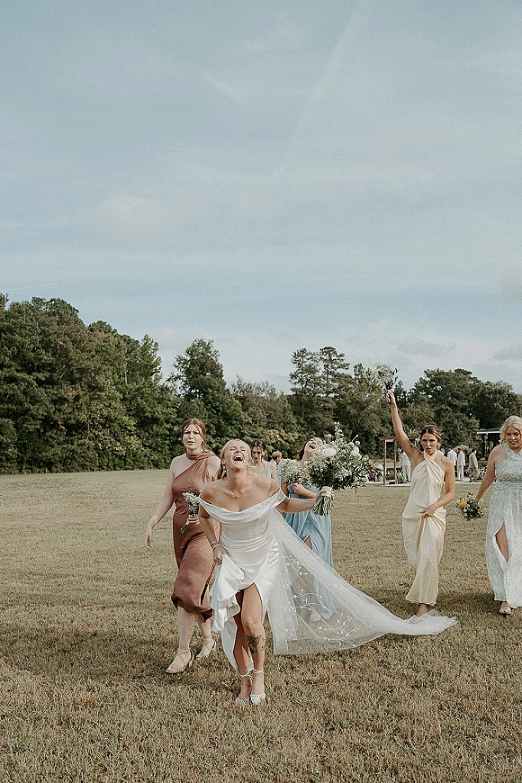 Bride with bridesmaids walking and laughing, holding bouquets in neutral dresses beside a grassy field with trees under a cloudy sky