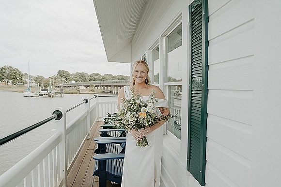 Bridal portrait of a bride holding bouquet in an off the shoulder wedding dress on a porch overlooking a waterfront dock and boats