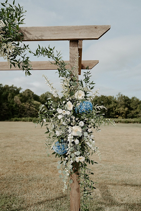 Wedding altar flowers on a wood wedding altar with eucalyptus greenery and white blooms, blue hydrangea, set in an open field with trees and sky