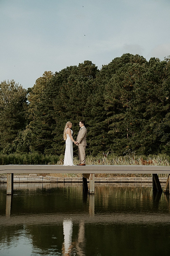 Couple portrait of bride and groom holding hands on a wooden dock, her half-up hair and sheath dress framed by pond reflections and treeline