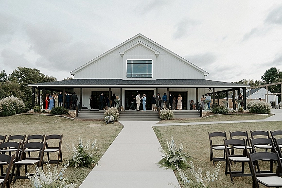 Outdoor ceremony setup with barn wedding ceremony aisle runner, black chairs and greenery florals leading to a white barn porch under string lights