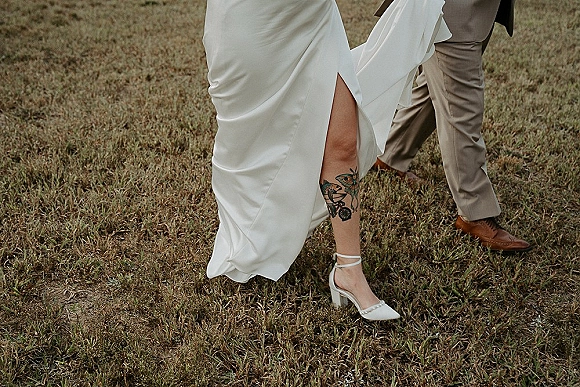 Bride shoe detail with wedding heel close up as ankle strap heels step from a slit white dress beside groom’s brown shoes on grass field
