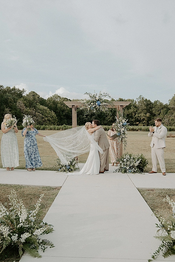 Ceremony kiss at an outdoor wedding ceremony as the bride and groom kiss under a wooden wedding arch in an open field of trees and sky