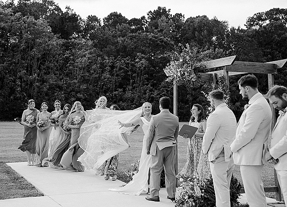 Ceremony moment as bride and groom at altar beneath a floral arch, veil blowing as bridesmaids and groomsmen line a grassy lawn