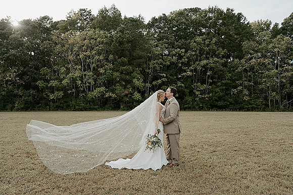 Wedding couple portrait of bride and groom kiss as her long cathedral veil blows across an open field with a distant tree line