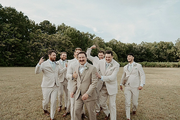 Groomsmen portrait of the groom with groomsmen in beige suits and boutonnieres, walking across a grassy open field with tree line and sky behind