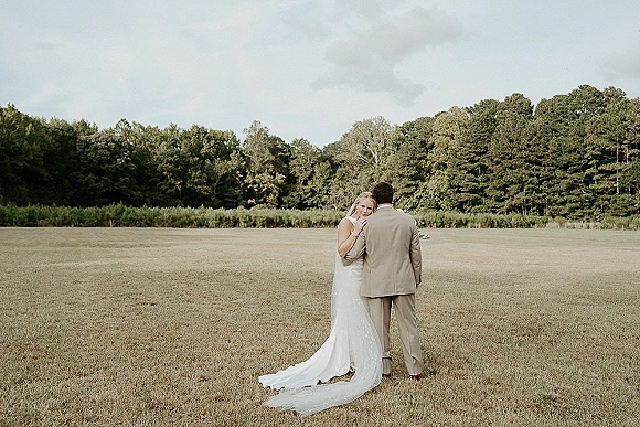 Couple portrait of bride and groom embrace in an open field, her long wedding dress train and veil flowing behind under a cloudy sky