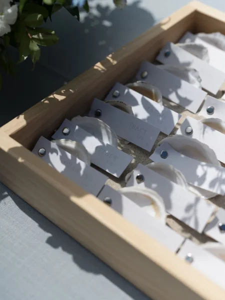 Escort cards on oyster shells with white calligraphy place cards, metal rivets, sand and greenery in a wooden tray on blue tablecloth