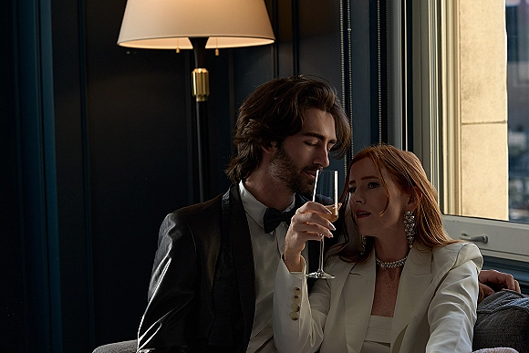 Couple portrait of newlyweds on a sofa, groom in tuxedo whispering as the bride in a white blazer holds a champagne flute by a window city view