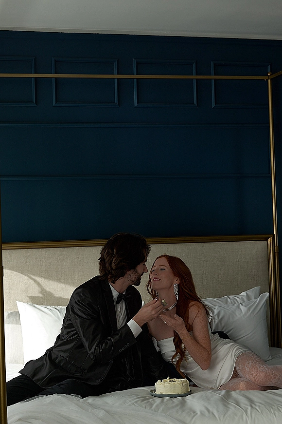 Couple portrait of bride and groom on bed, groom feeding bride wedding cake beside pillows, with navy paneled wall and canopy frame