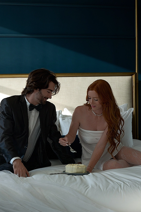 Wedding couple portrait of bride and groom on bed cutting wedding cake, she in a slip dress with pearl necklace, blue paneled bedroom behind