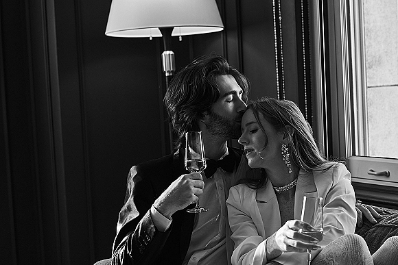Couple portrait of newlyweds in a tuxedo and white blazer holding champagne flutes, sharing a forehead kiss on a sofa by window light