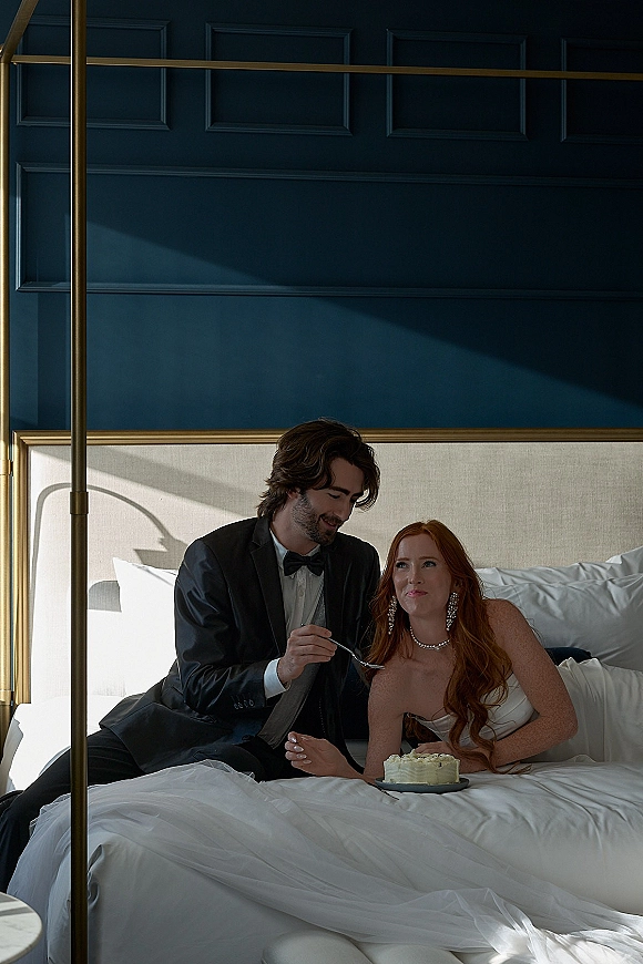 Couple portrait of bride and groom on bed, sharing a bite of wedding cake in a sunlit bedroom with paneled wall shadows