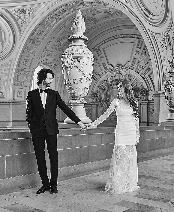 Couple portrait of bride and groom holding hands in formal attire on stone steps beneath ornate arches and carved ceiling, black and white