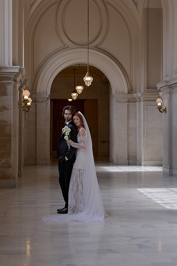 Couple portrait of bride and groom posing, her lace-sleeve dress and long veil with bouquet, in a grand arched marble hallway