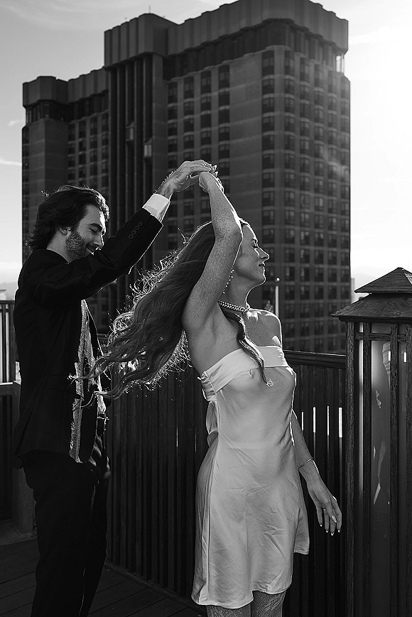 Couple portrait of groom spinning the bride in a strapless wedding dress twirl on a rooftop deck, with wooden railing and high-rise sky backdrop