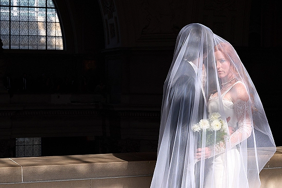 Couple portrait of bride and groom under veil, bride holding bouquet, posed by an arched window with dramatic indoor shadows
