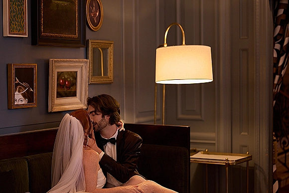 Wedding kiss portrait of bride and groom kissing on a sofa, her long veil and strapless dress beside a floor lamp and framed art wall