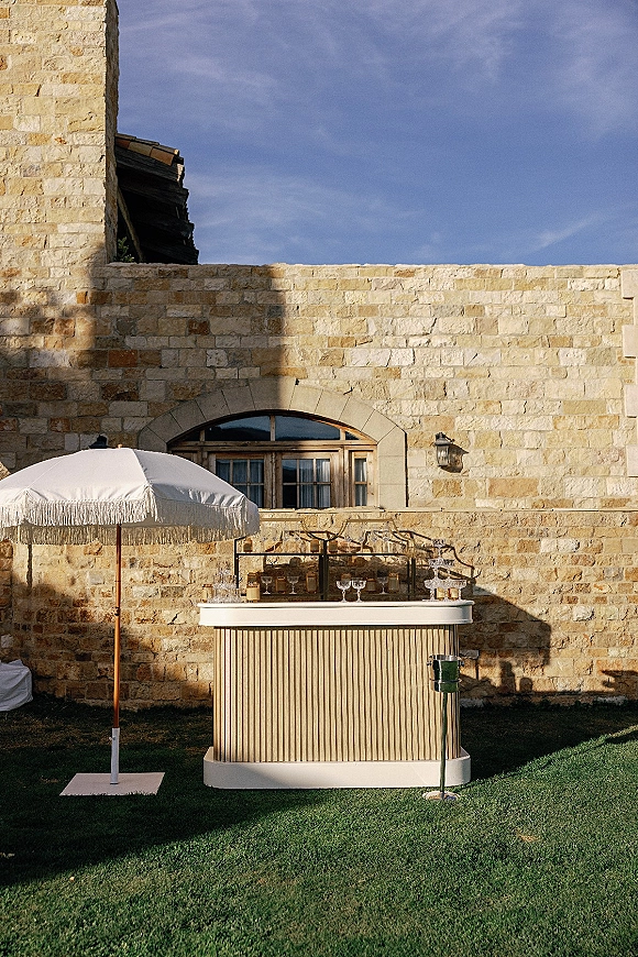 Wedding bar setup with mobile bar under a fringe umbrella, coupe glasses and bottles displayed on shelves beside a stone wall window