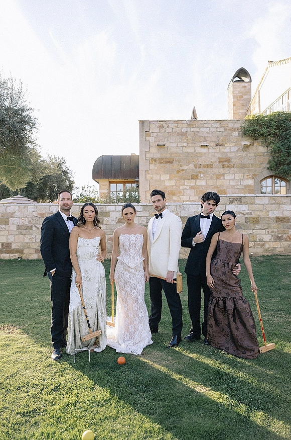 Wedding party portrait of bride and groom with friends in black tie attire holding croquet mallets on a lawn by a stone building