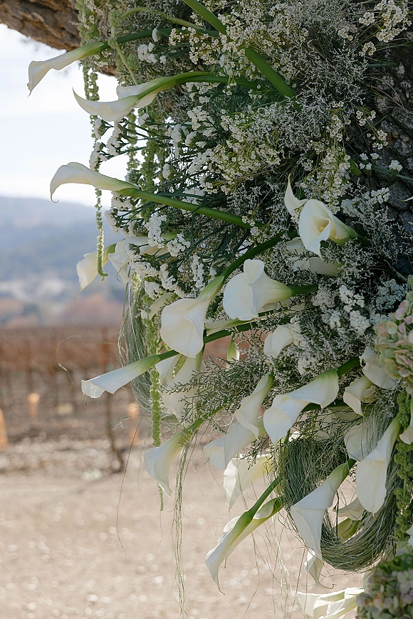 Wedding floral arch with calla lily arch flowers and hanging greenery, asymmetrical white blooms against hills, bare trees, and sky