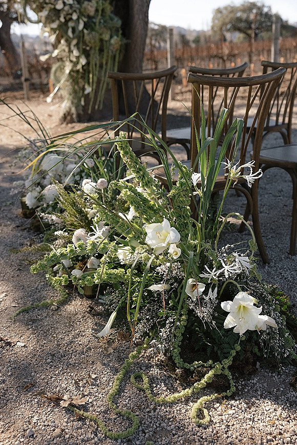 Ceremony aisle florals with grounded aisle flowers of white lilies, roses and greenery lining a gravel path between wooden chairs in a vineyard setting
