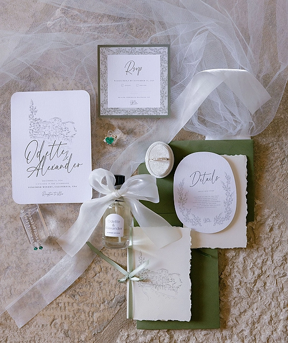 Invitation suite wedding invitation flat lay with vellum wrap and satin bow, olive envelopes, RSVP card, ring and perfume on stone surface