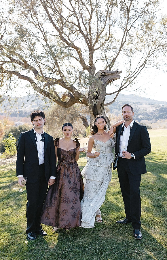 Wedding party portrait with bridesmaid and groomsmen photo, all holding champagne flutes on a sunny lawn under a large tree