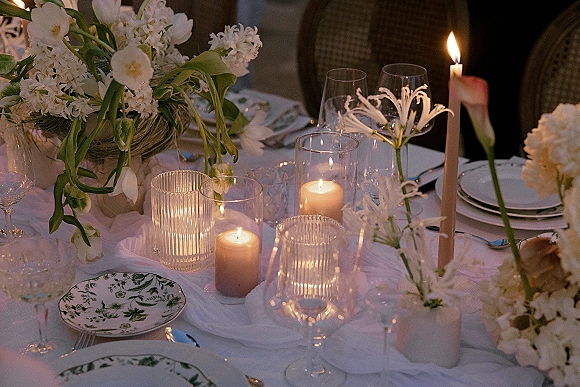 Reception tablescape with wedding table decor featuring white florals and taper candles in ribbed glass holders on white linen in dim light