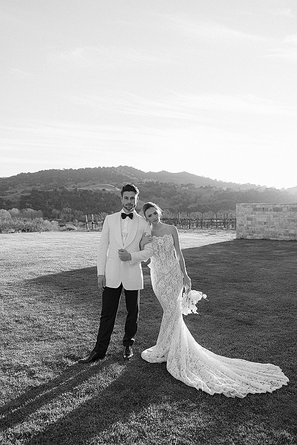 Couple portrait in a black and white wedding portrait, bride in strapless lace gown with bouquet leaning on groom by vineyard rows