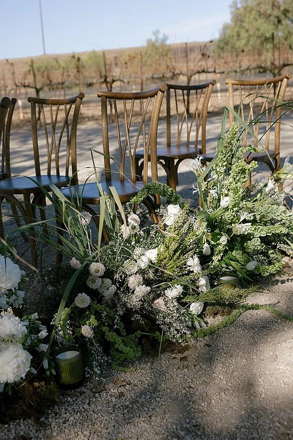 Ceremony aisle decor with a ground floral aisle of white blooms and greenery, with votive candles along a gravel path in a vineyard