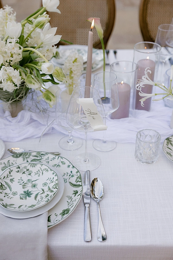 Reception tablescape with a wedding place setting, white floral centerpiece, taper candles in glass hurricanes, and place card on a wine glass