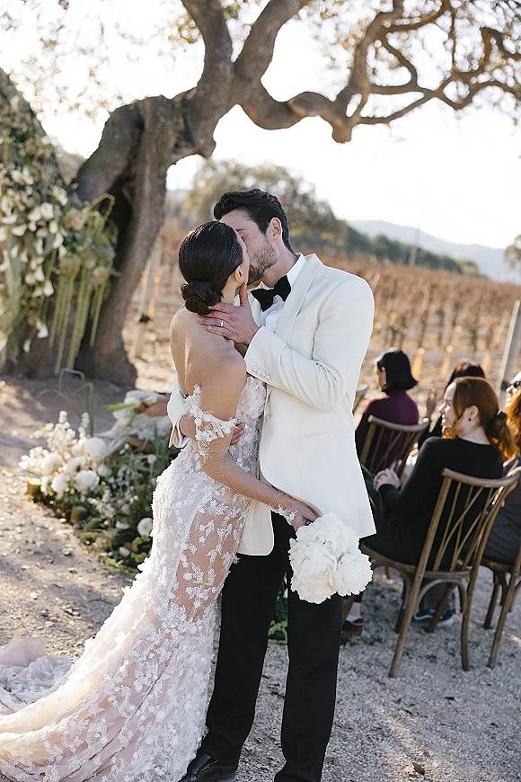 Wedding kiss as bride in lace gown with long train embraces groom in white tuxedo under oak tree, vineyard rows and mountains behind