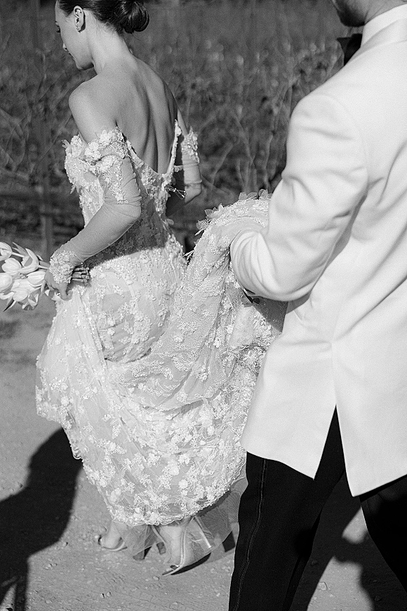 Wedding couple portrait of bride and groom walking away, groom holding dress train on an outdoor path through a tall grass field