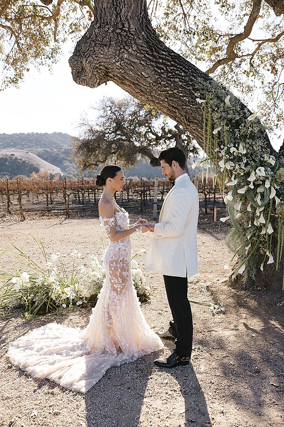 Ceremony moment of couple exchanging vows under a large oak tree, bride in off-shoulder lace gown with train, calla lilies and greenery backdrop