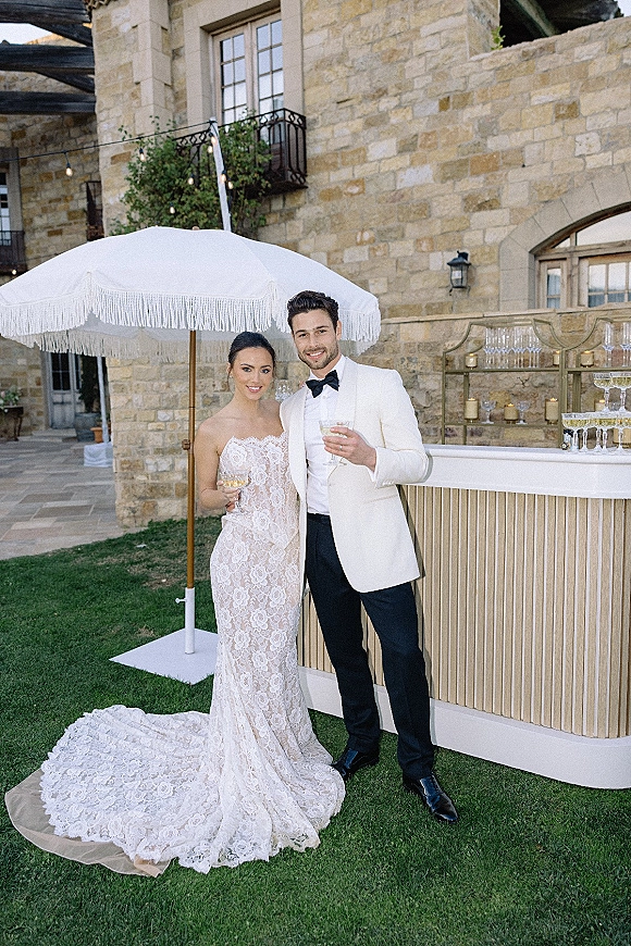 Couple portrait of bride and groom with champagne, her lace train and his white tux, toasting by an outdoor bar with candlelit tower
