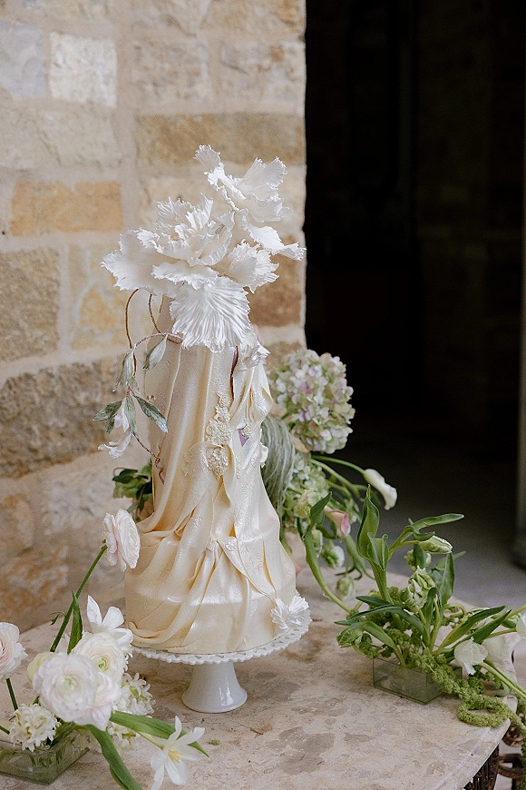 Wedding cake with textured buttercream, draped fabric, and white flowers with greenery on a pedestal atop a stone table by a stone wall