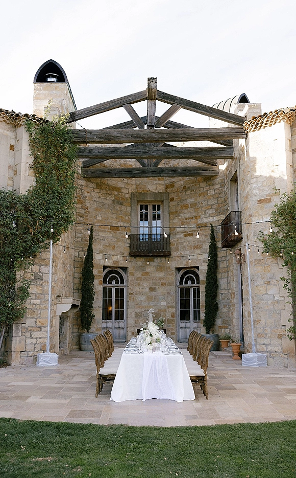 Reception tablescape with long banquet table wedding setup, white tablecloth, wicker chairs, white floral centerpiece, string lights in stone courtyard